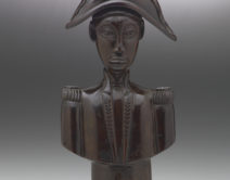 A wooden, sculpted bust of Jean-Jaques Dessaline, he is dressed in military uniform with a feathered, bicorne hat. His face is quite narrow and stylised.